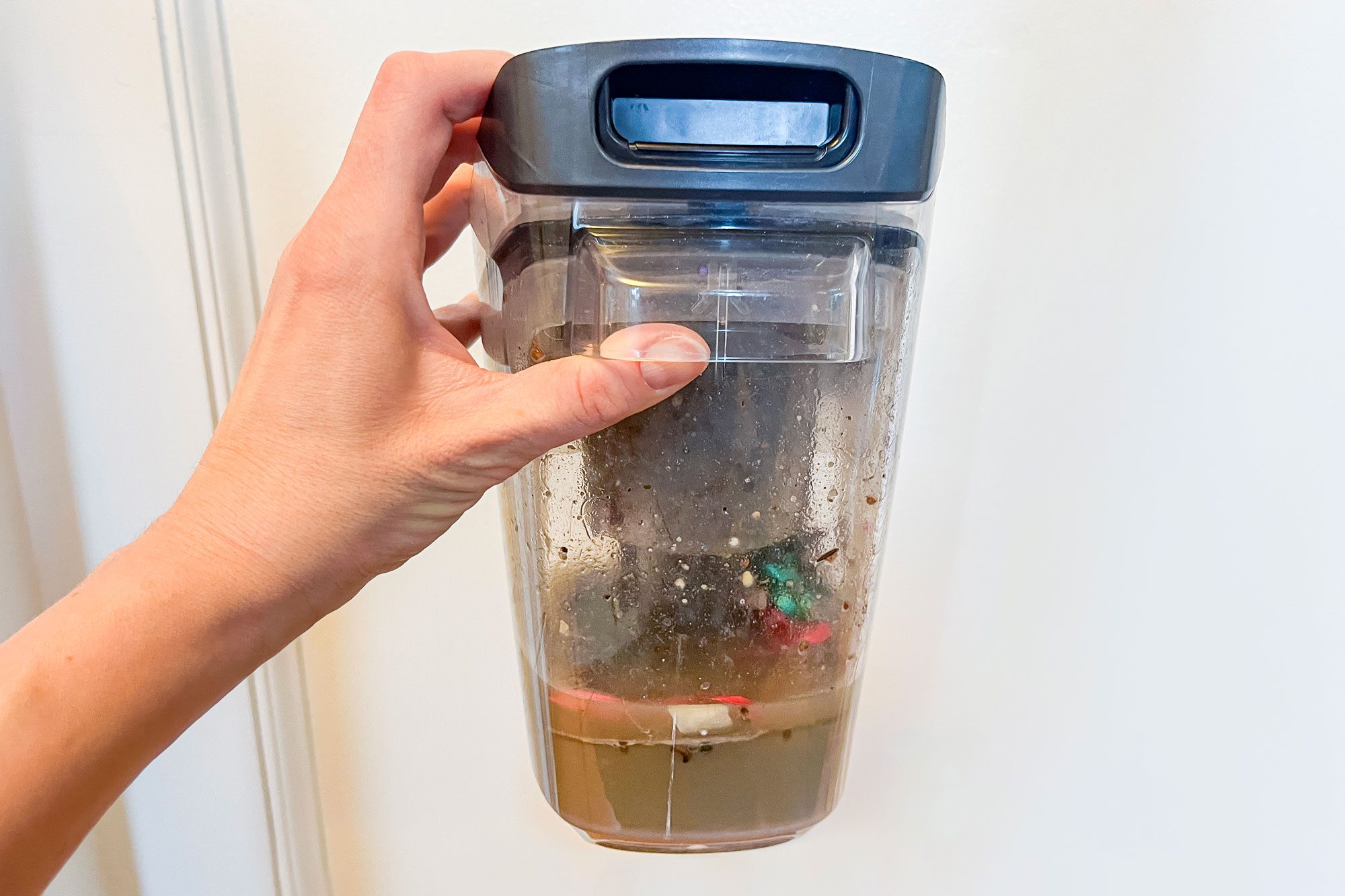 Hand holds a clear, dirty vacuum cleaner canister against a plain wall, showcasing collected debris and liquid inside.