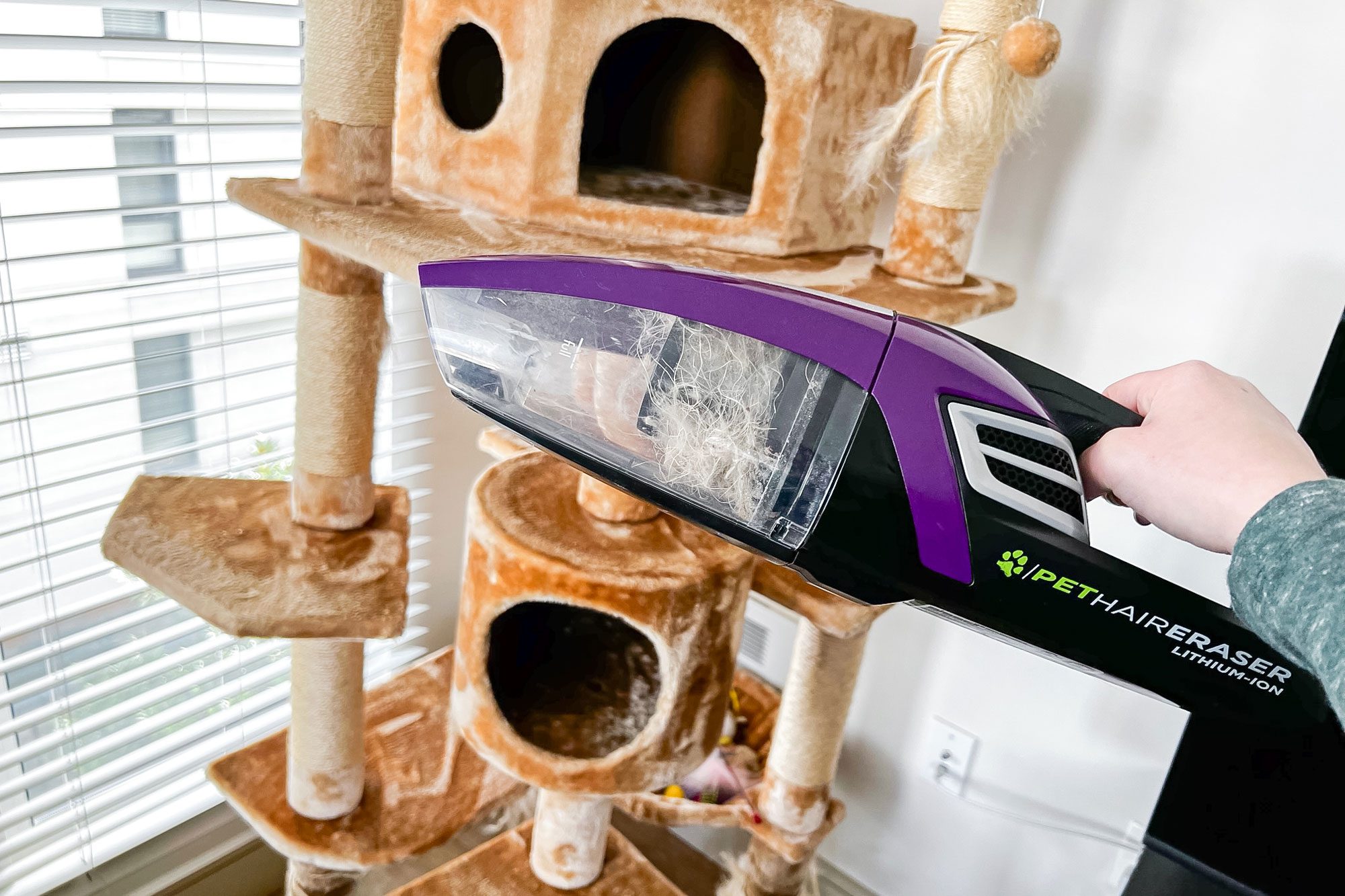 Handheld vacuum cleaning cat tree, collecting fur; sunlight filters through blinds onto plush structure. "PET HAIR ERASER Lithium-Ion" is visible on vacuum.