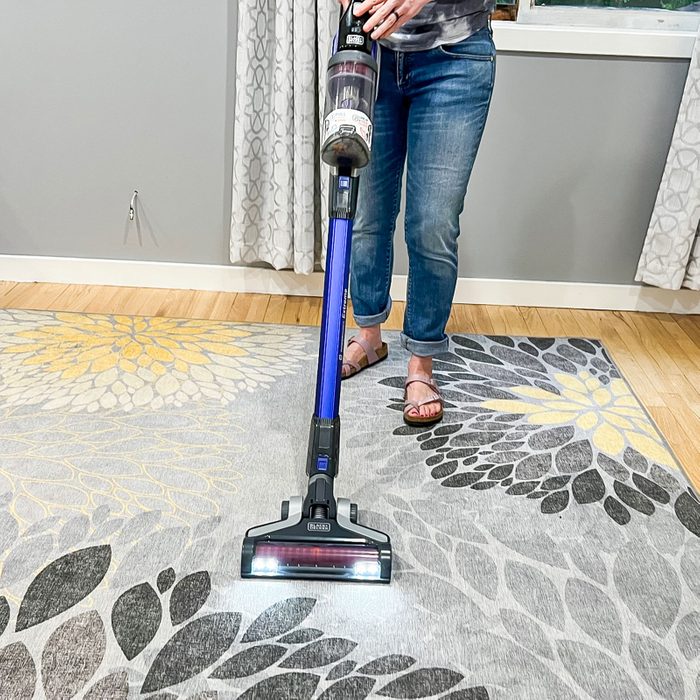 Vacuum cleans gray floral-patterned rug in a room with wooden floor and gray curtains, while a person in jeans and sandals operates it.