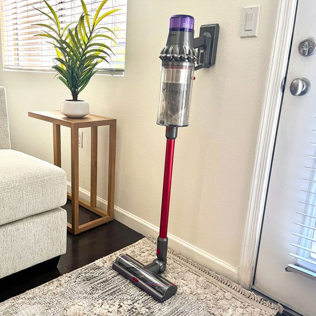 Cordless vacuum cleaner rests on the wall, near a beige sofa and wooden side table with a potted plant, in a sunlit room.