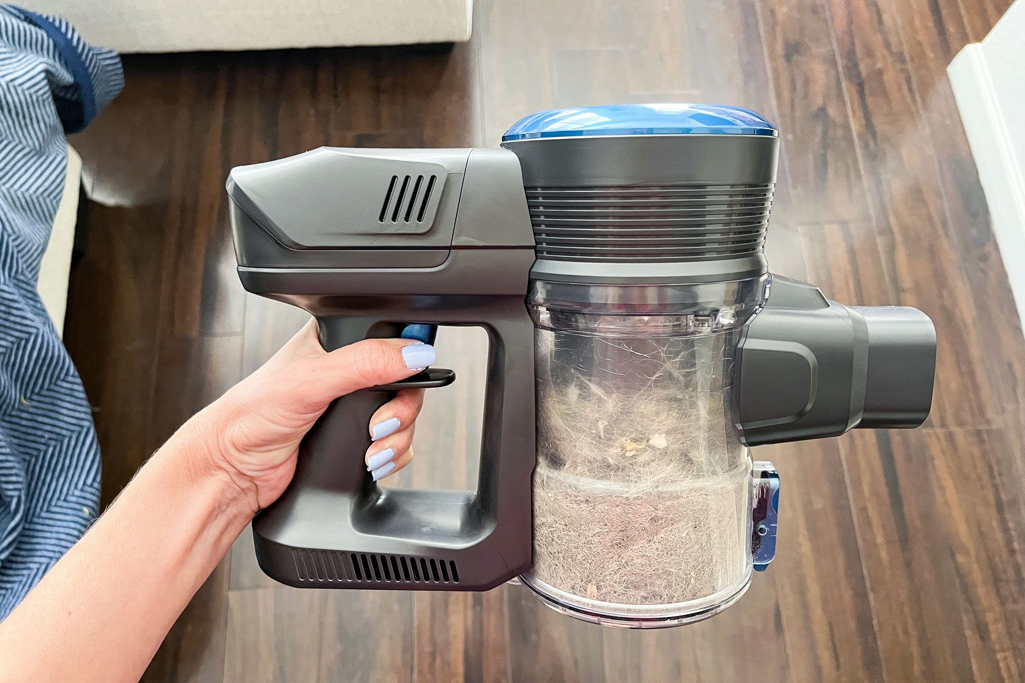 Hand holds cordless vacuum filled with dust, against a wooden floor and partially visible striped fabric.