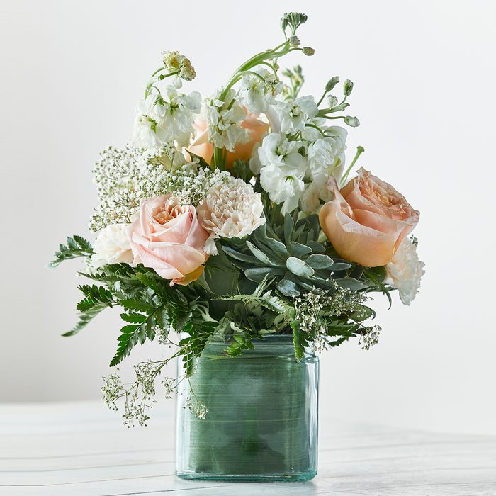 Flowers arranged in a glass vase sit elegantly, featuring roses and greenery against a white background on a light wooden surface.