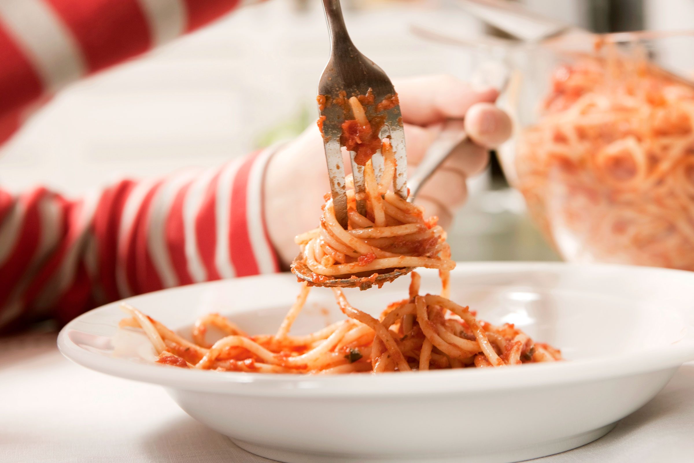 Fork twirls spaghetti on a plate, with a red-striped sleeve visible, set at a dining table with a glass bowl of pasta nearby.