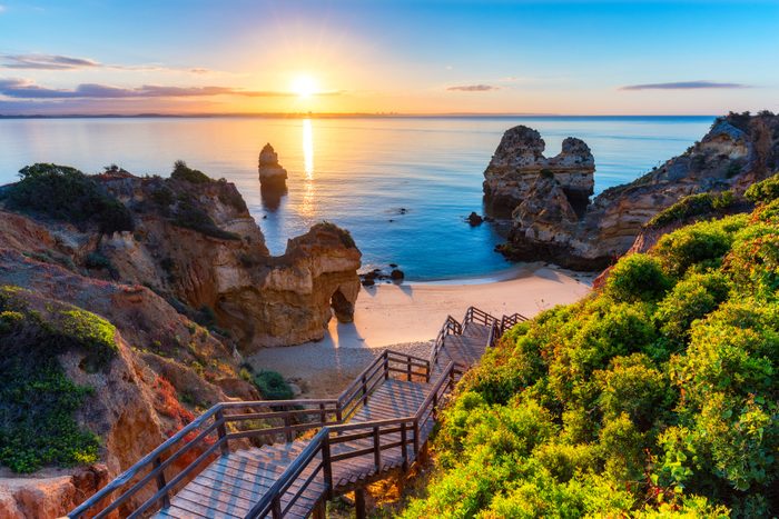 Wooden staircase descends towards serene beach; rocky formations and calm sea illuminated by sunset in vibrant coastal landscape.