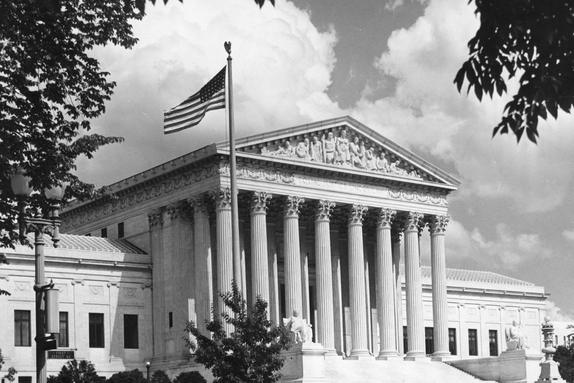 The classical building stands adorned with columns, an American flag waving above, surrounded by trees and under a partly cloudy sky.