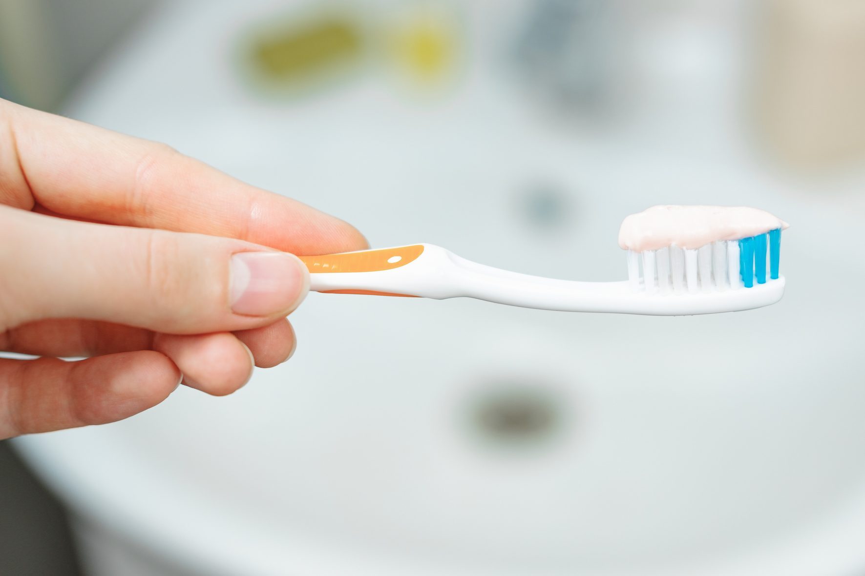 Toothbrush, held horizontally, with pink toothpaste on bristles; background shows a blurred bathroom sink.