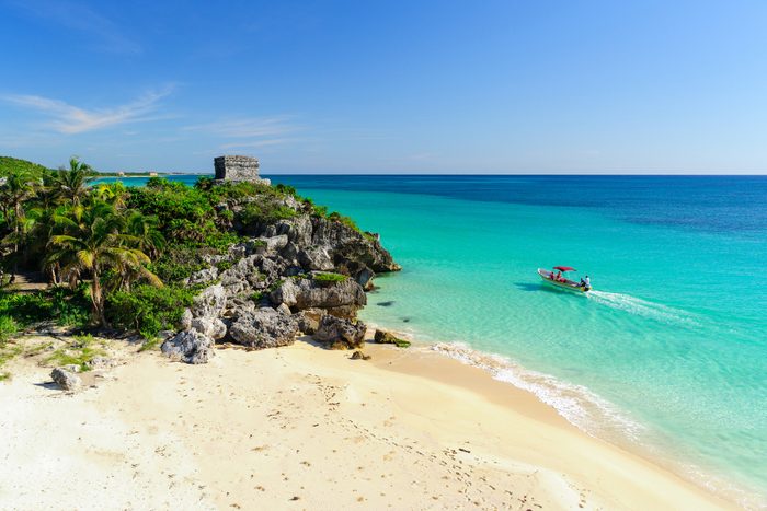 A small boat navigates crystal-clear turquoise waters near a sandy beach with rocky cliffs, surrounded by lush greenery and a bright blue sky.