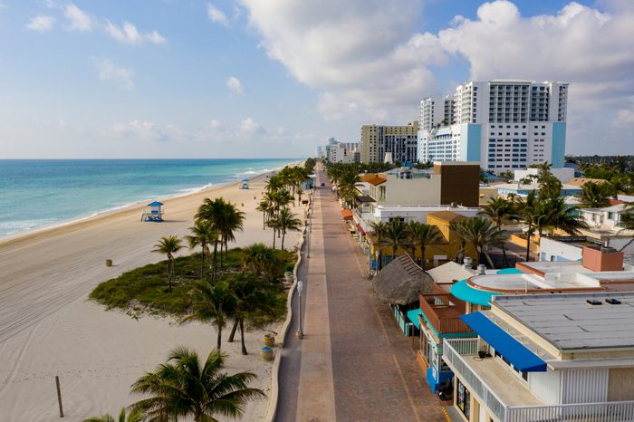 Palm trees line a beachside promenade, bordered by colorful buildings and high-rises, under a partly cloudy sky with turquoise ocean in the background.