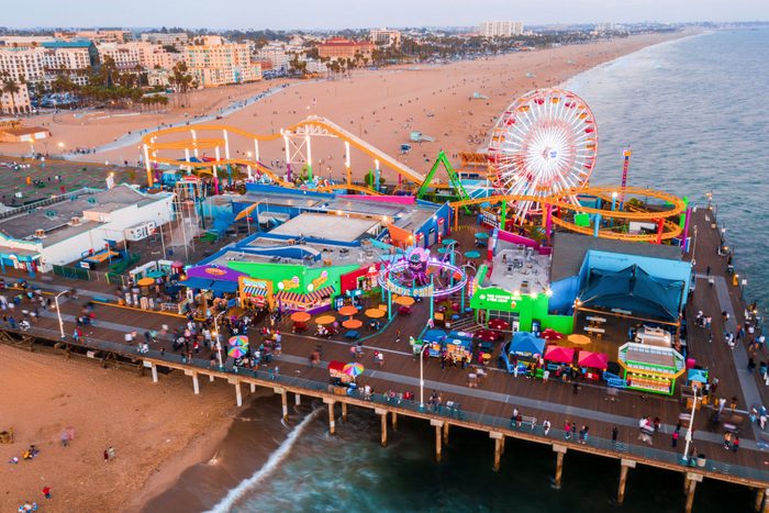 Ferris wheel spins on a colorful amusement pier, next to the ocean. Beachgoers and visitors explore the lively boardwalk, with city buildings in the background.