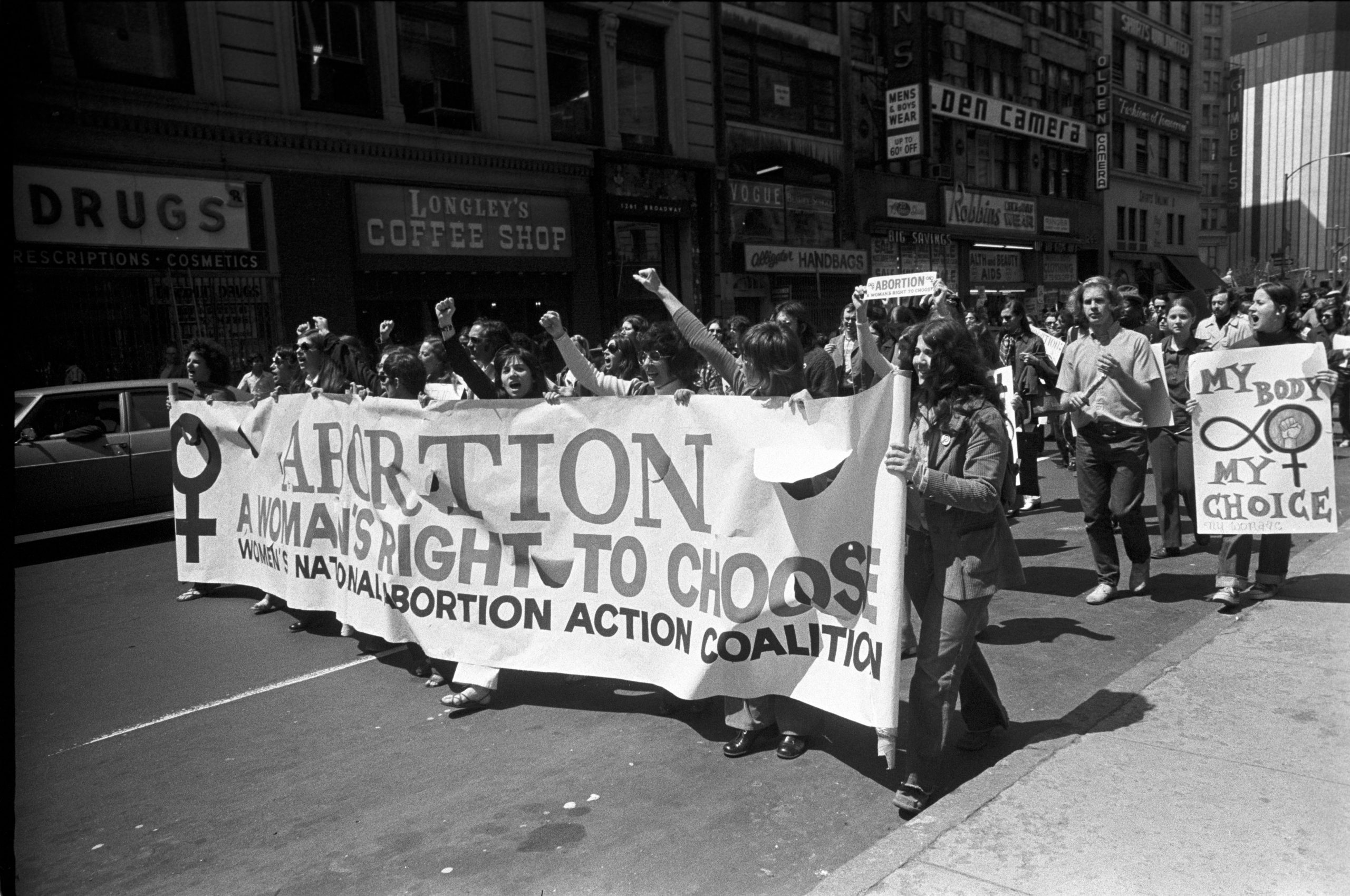 Protesters march, holding a large banner reading "ABORTION A WOMAN'S RIGHT TO CHOOSE," on a city street surrounded by storefronts.