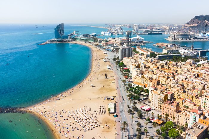 Beach curves alongside city buildings, with people lounging under umbrellas; clear blue sea and a distinctive, sail-shaped hotel in the background.