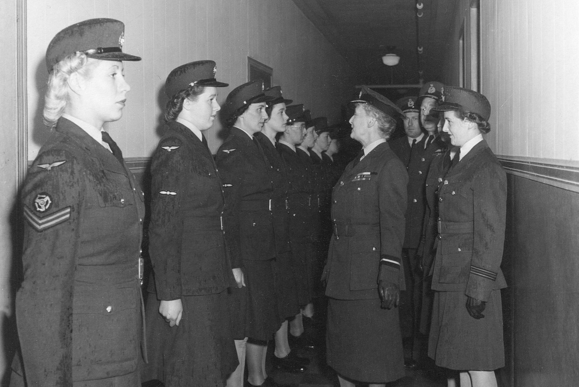 Uniformed women stand in formation, facing each other, while being inspected by a senior officer, inside a narrow corridor.
