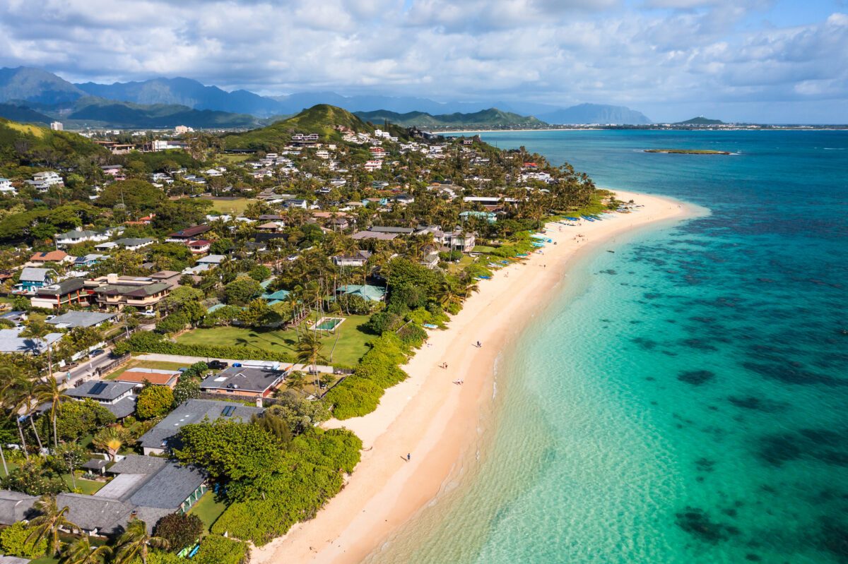 Beach stretches along a coastal town with turquoise waters and scattered houses, backed by lush green hills and blue mountains under a partly cloudy sky.