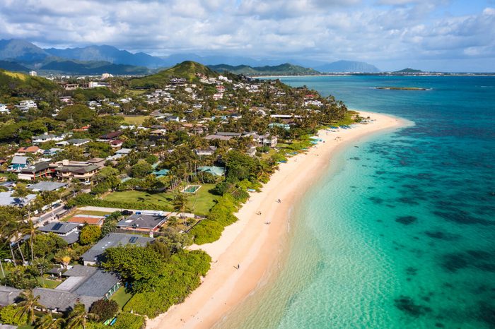 Beach stretches along a coastal town with turquoise waters and scattered houses, backed by lush green hills and blue mountains under a partly cloudy sky.