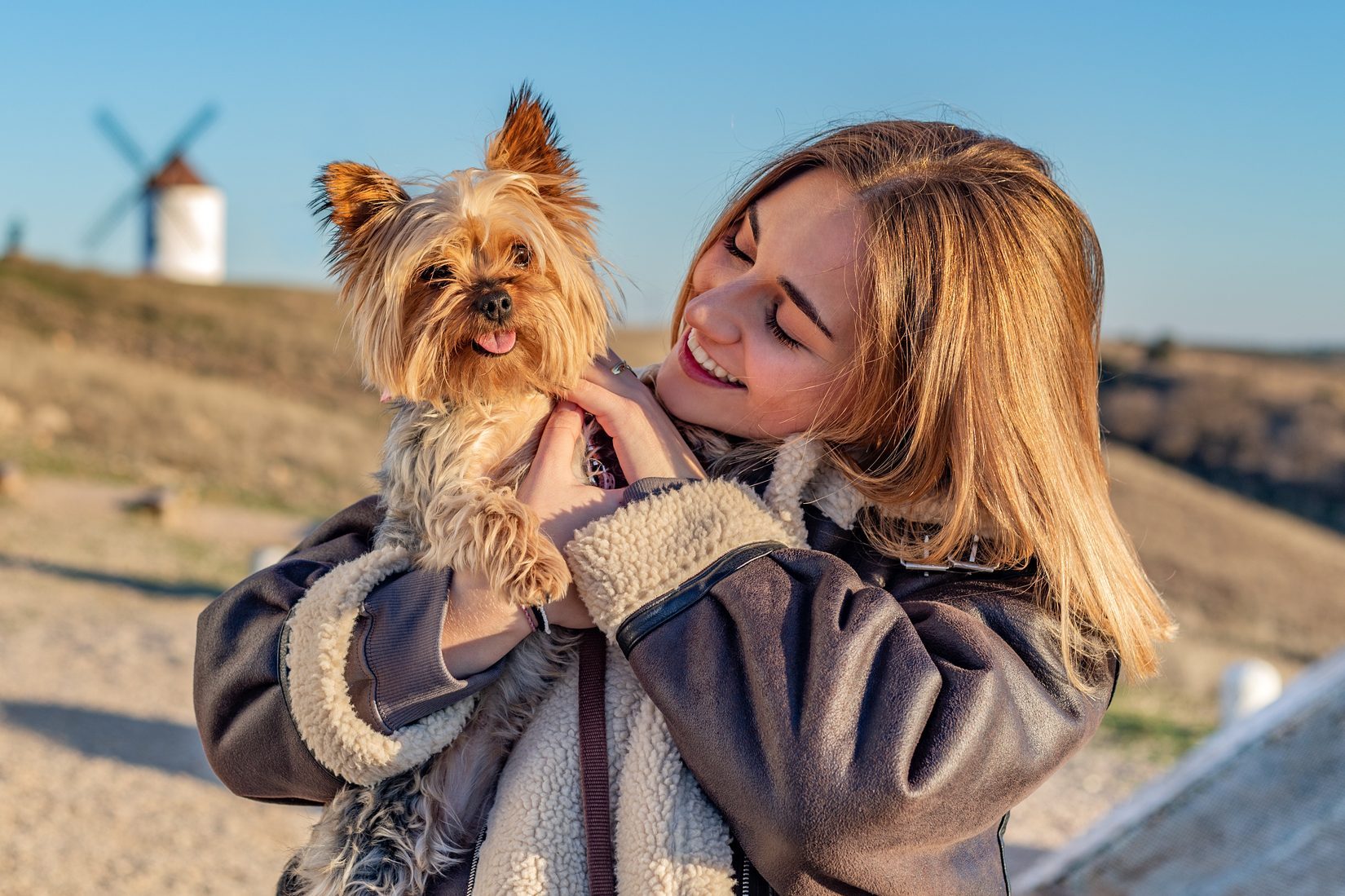 A small dog is held by a smiling woman outdoors, with a windmill and a clear blue sky in the background.