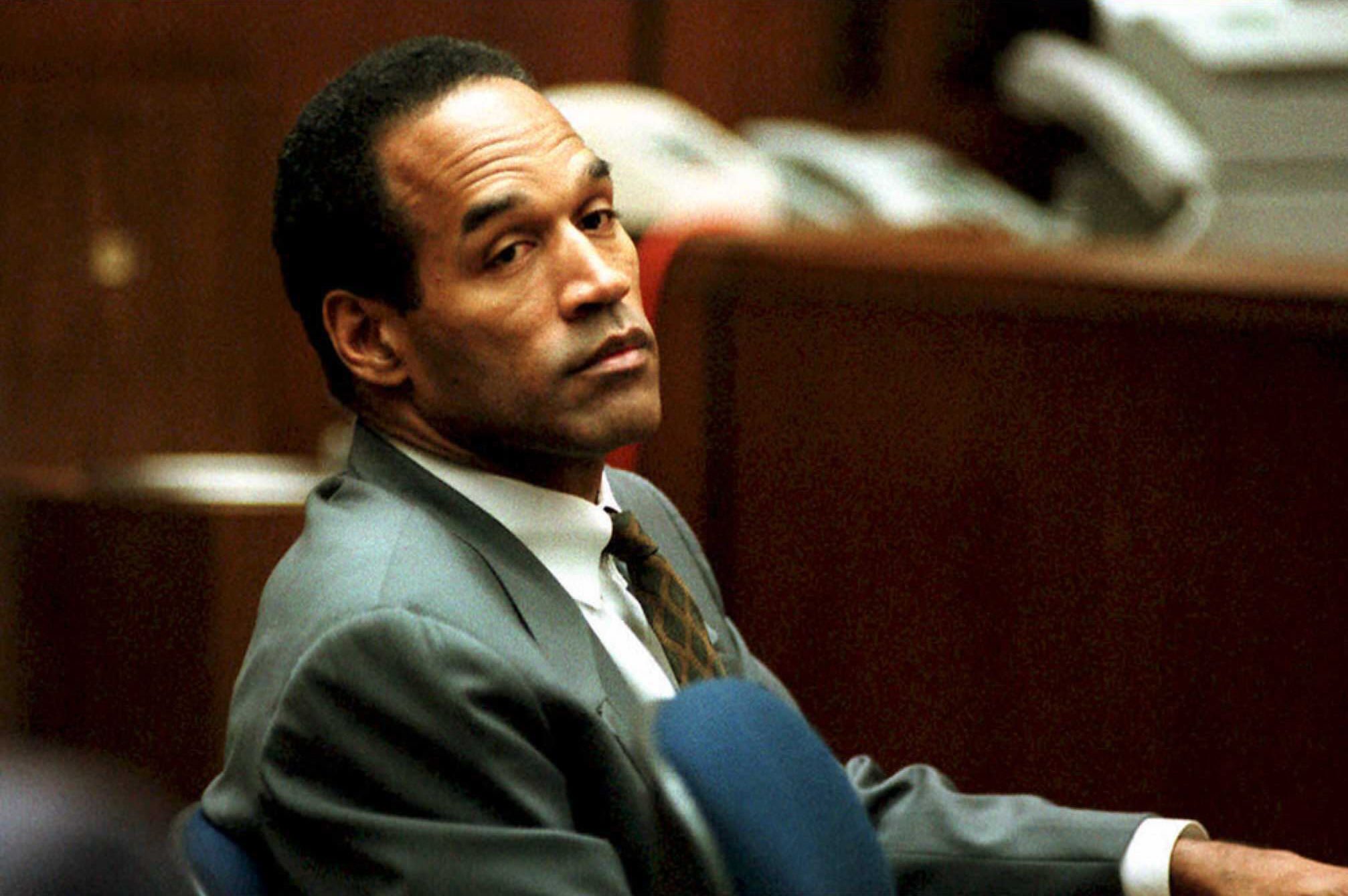 A person in a suit sits attentively in a courtroom setting, with blurred stacks of books and telephones in the background.