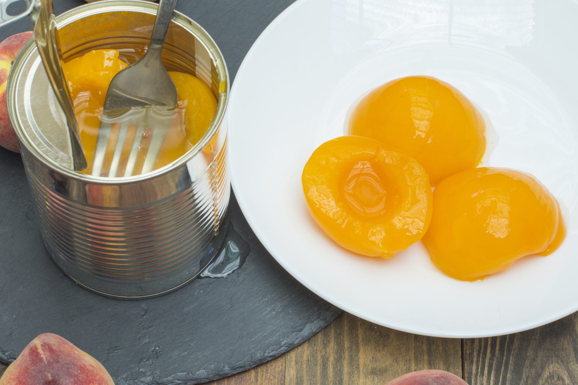 Canned peaches sit in syrup with a fork, next to a white plate holding peach halves, surrounded by fresh peaches on a slate board.