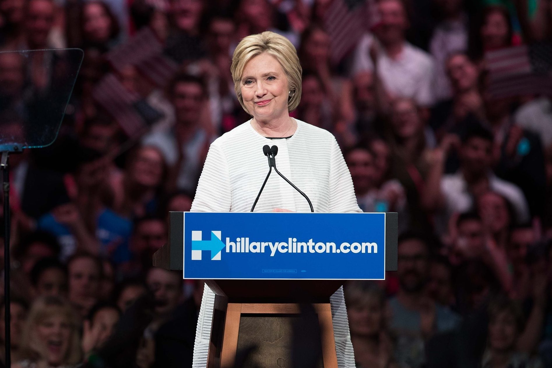 A person speaks at a podium displaying "hillaryclinton.com" against a backdrop of a cheering crowd with American flags.