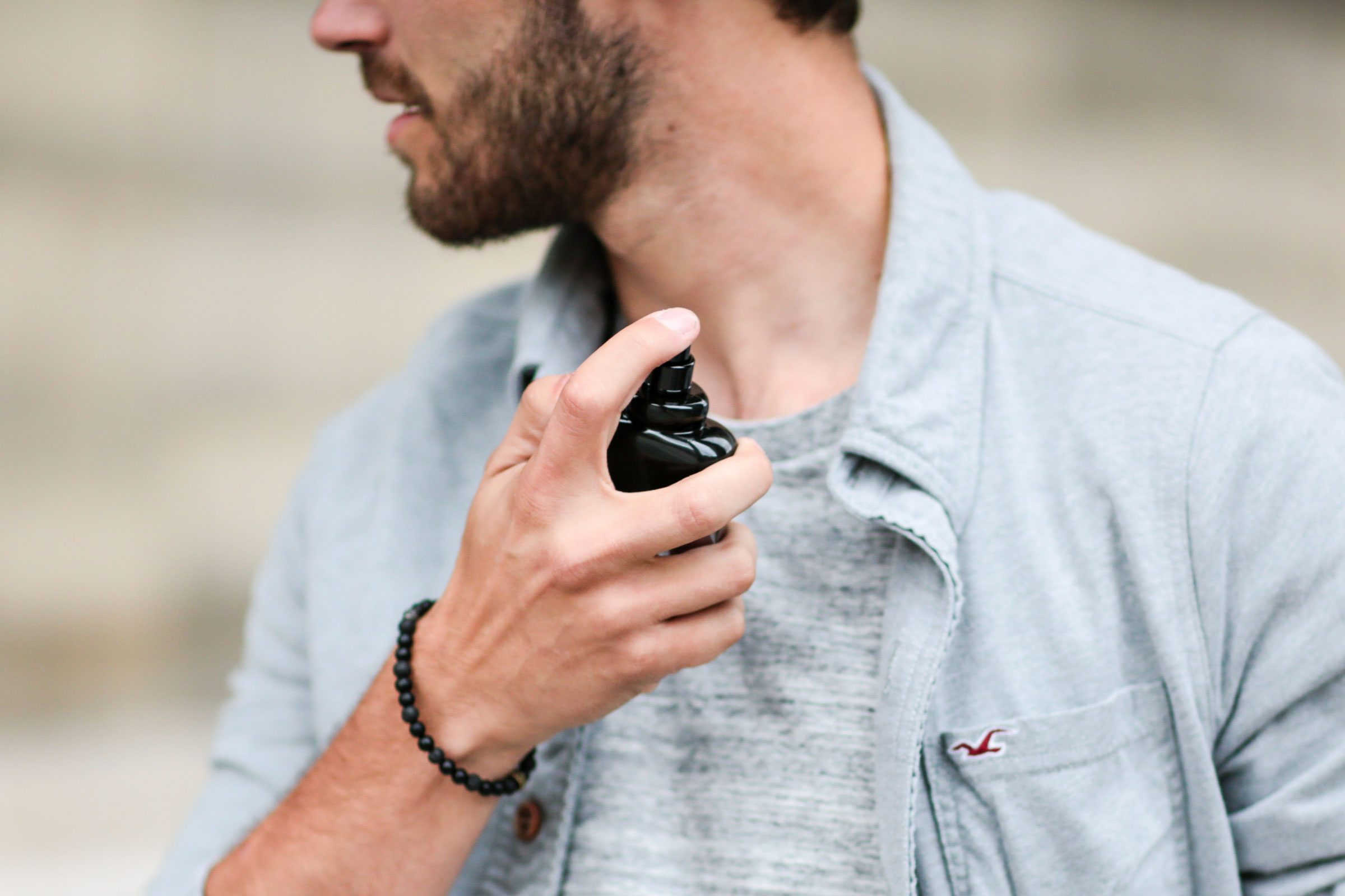 A person sprays cologne on their neck while wearing a light gray shirt and beaded bracelet, against a blurred background.