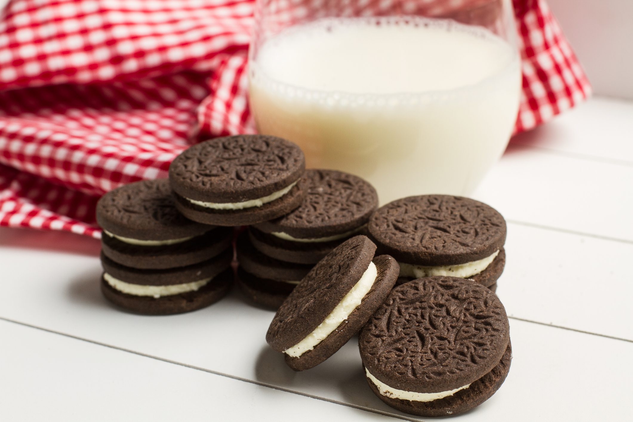 Chocolate sandwich cookies are stacked beside a glass of milk on a white table, with a red and white checkered cloth nearby.
