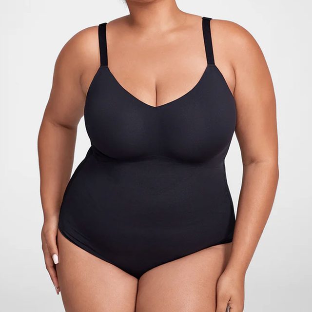 A person stands wearing a black bodysuit, shown from shoulders to thighs, in front of a plain, light-gray background.