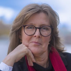 A person with glasses rests their chin on their hand, wearing a red scarf, with a blurred outdoor background.
