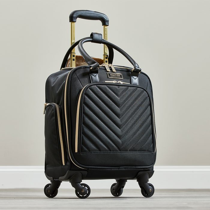 Black rolling suitcase stands on a wooden floor, featuring gold accents and a chevron-patterned front pocket.