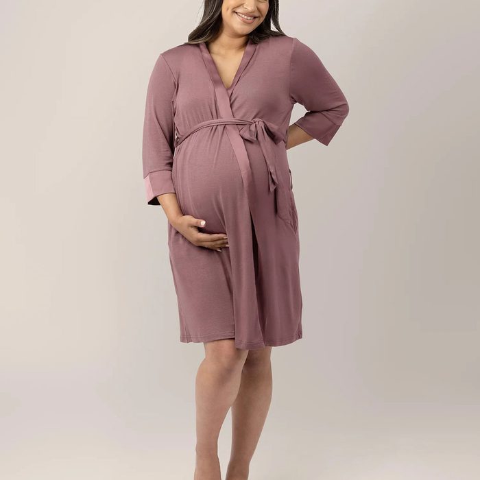 Person wearing mauve robe cradles pregnant belly, standing against a neutral background.