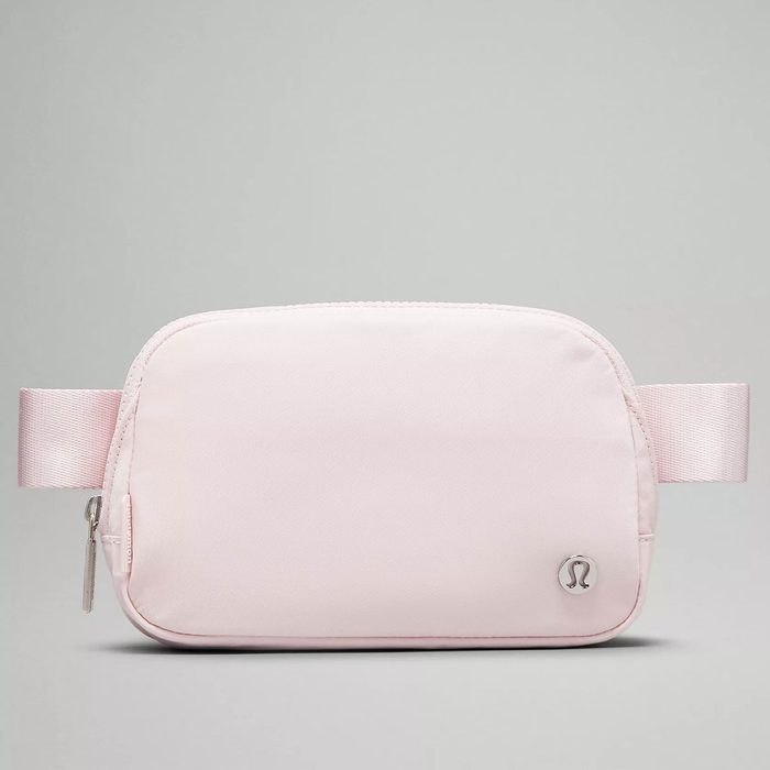 A light pink waist bag rests on a neutral background, featuring a zipper and a small metallic logo on the front.