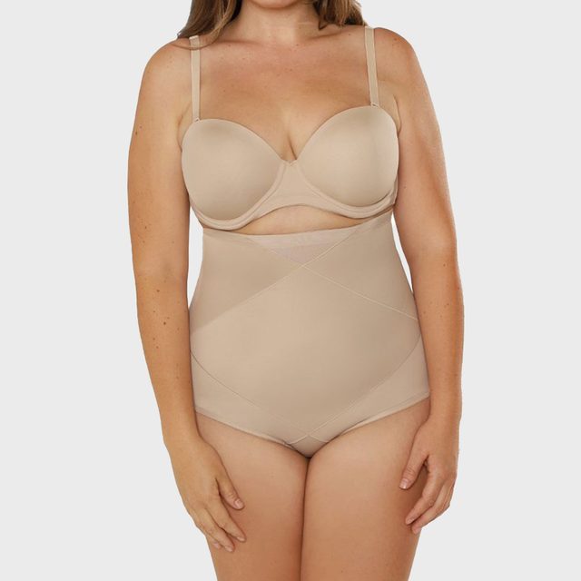A woman stands wearing beige shapewear and a bra against a plain white background.