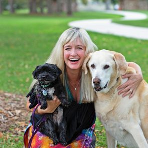 A person smiles while holding two dogs, a small black one and a larger yellow one, in a park with a winding path.