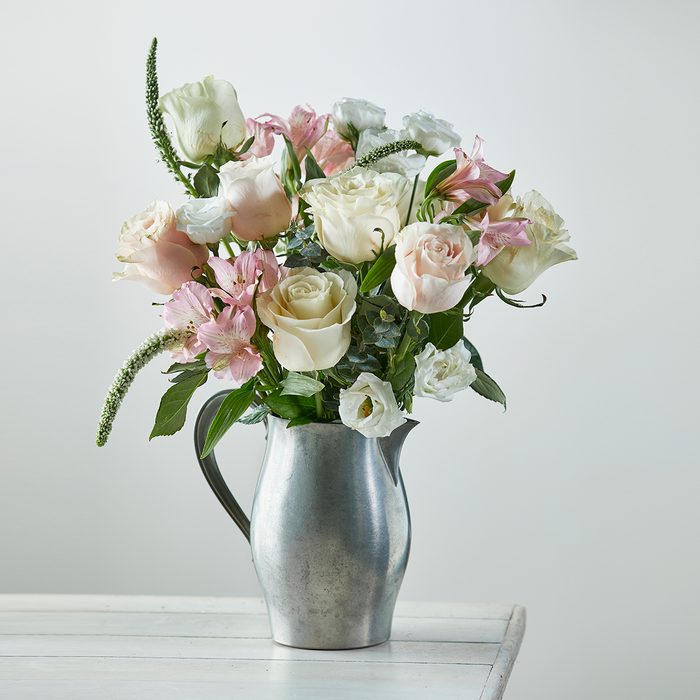 A metal pitcher holds pastel flowers, set on a white wooden table against a light background.