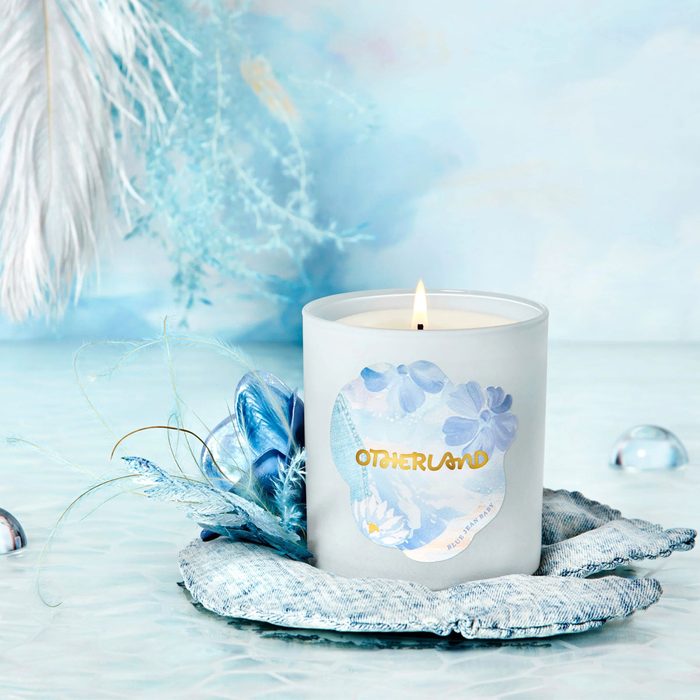 A white candle burns gently, surrounded by blue feathers, on a soft, textured surface in a serene, light blue setting. Text reads 