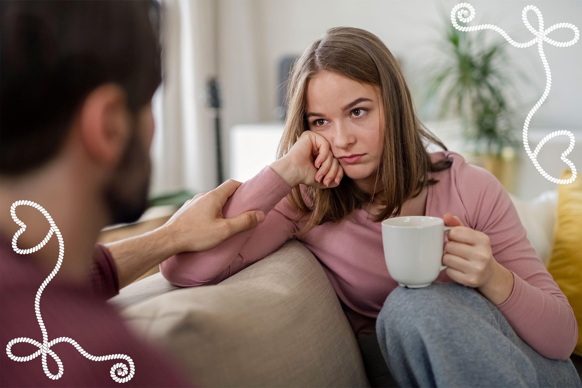 Woman holding mug looks contemplative; another person touches her arm. They're sitting on a couch in a cozy, plant-filled living room.
