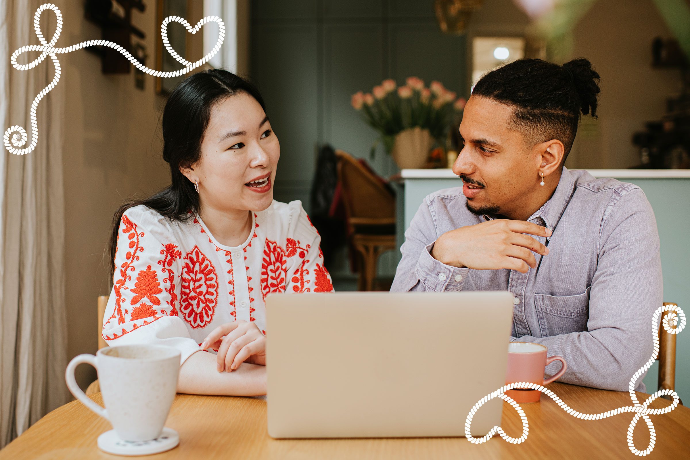 Laptop users converse over coffee at a wooden table, with floral decor in the background and decorative heart designs overlaying the image.