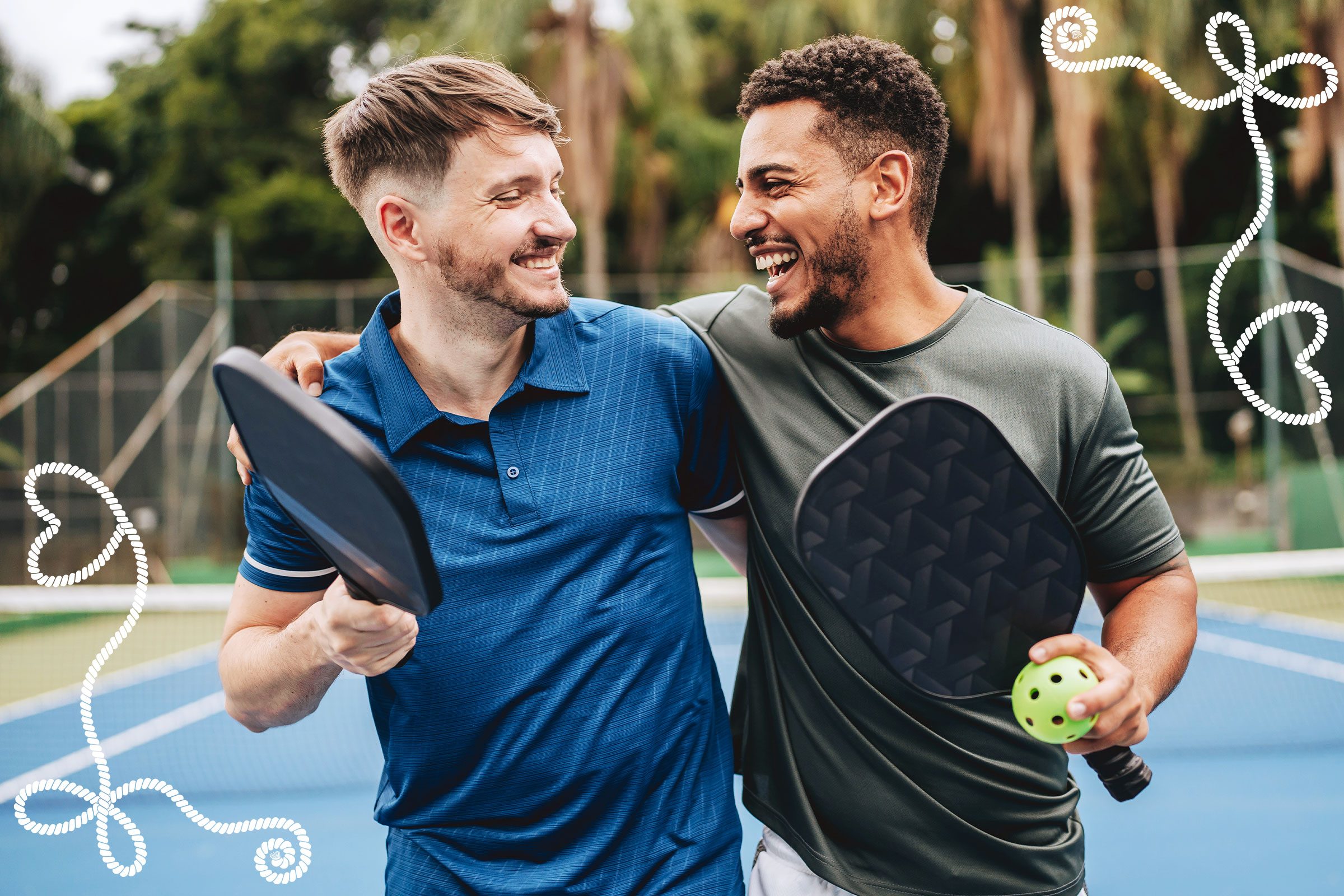 Two men smiling, holding pickleball paddles, on a tennis court with decorative white lines surrounding them.