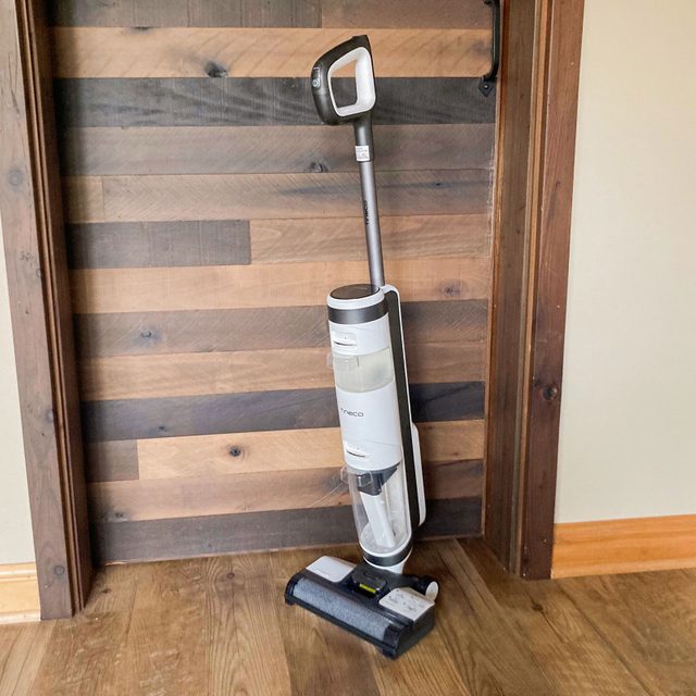 Vacuum cleaner standing upright against a wooden wall in a corner of a room with a hardwood floor.