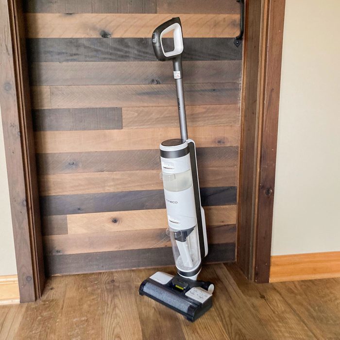 Vacuum cleaner standing upright against a wooden wall in a corner of a room with a hardwood floor.