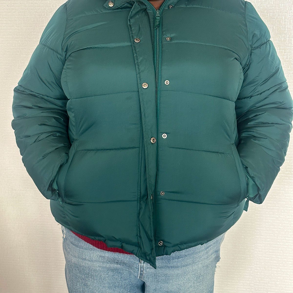 A person wears a buttoned green puffer jacket with hands in pockets, paired with blue jeans, against a textured light background.