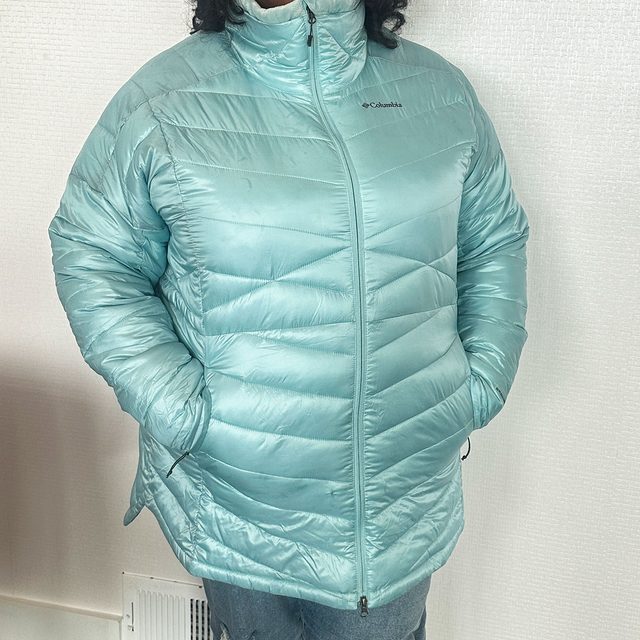 A person wears a light blue puffy jacket with hands in pockets, standing against a plain white textured wall.