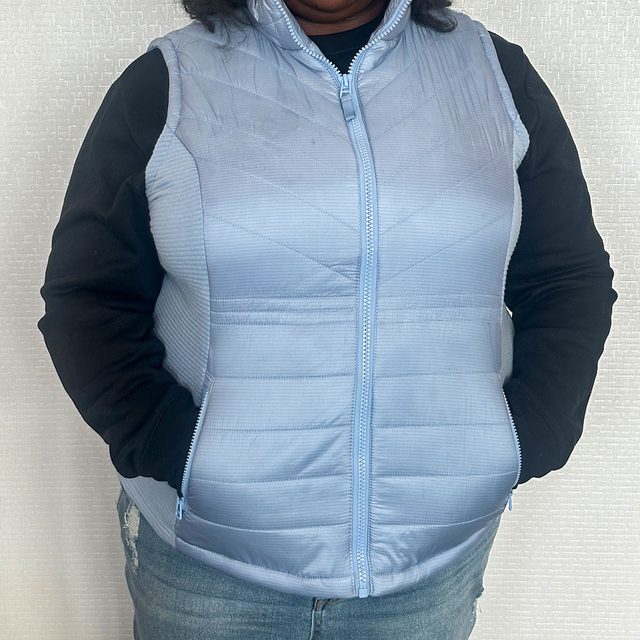 A person wears a light blue, quilted vest over a black long sleeve, standing against a textured light background.