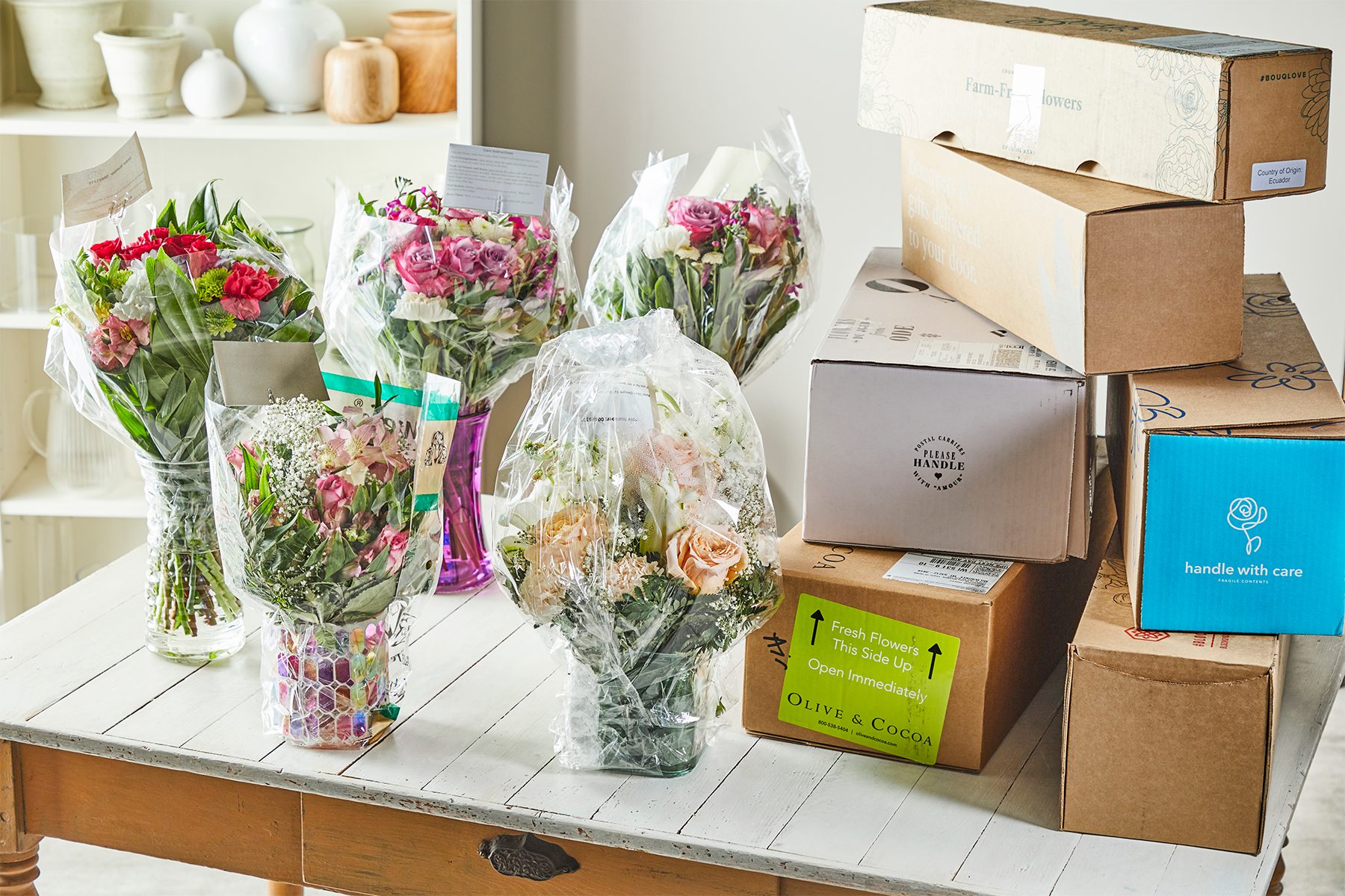 Flower bouquets stand in vases, surrounded by stacked cardboard boxes, on a wooden table in a bright room with shelves displaying various pots.
