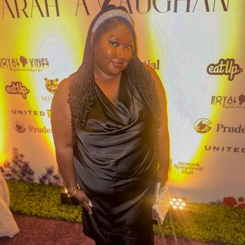 A person stands smiling wearing a black dress and headband, holding a sparkly clutch against a backdrop with logos and floral decorations.