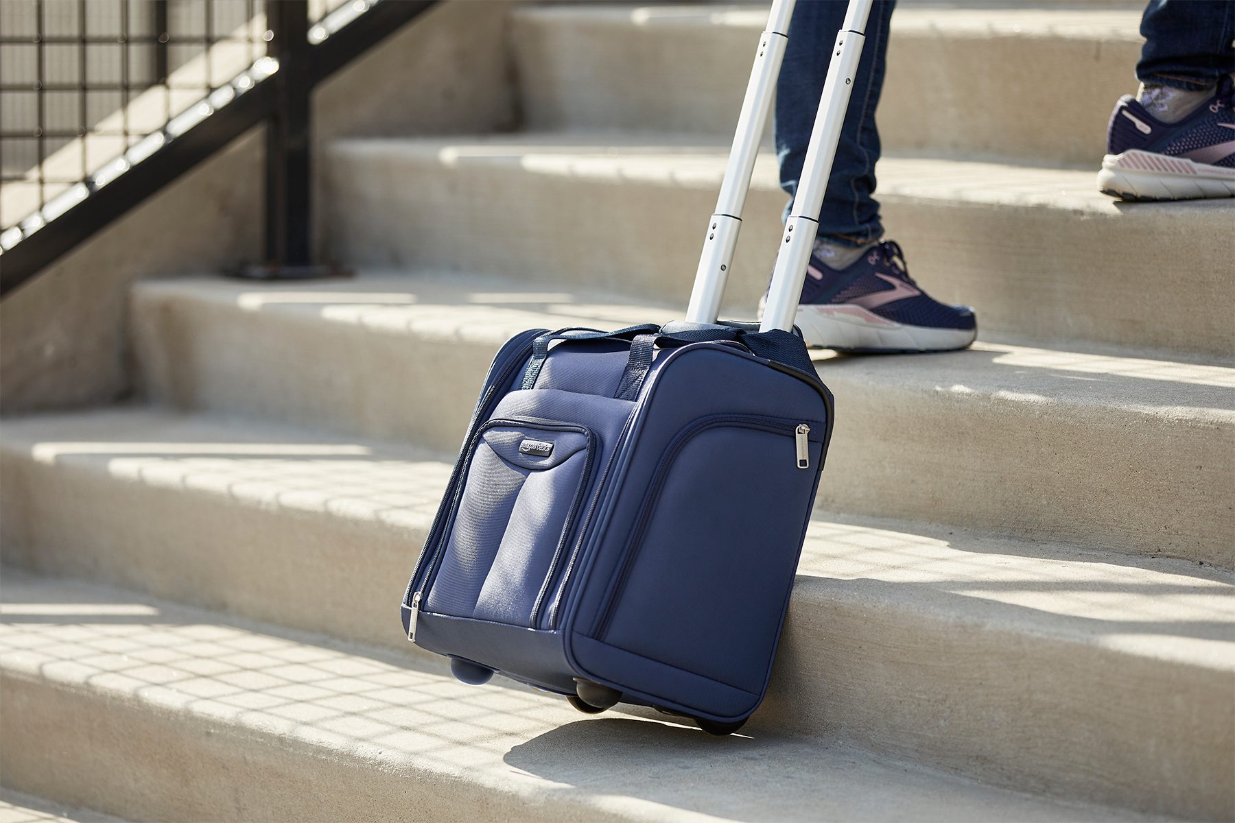 Blue wheeled suitcase being pulled up concrete stairs beside a person in sneakers.