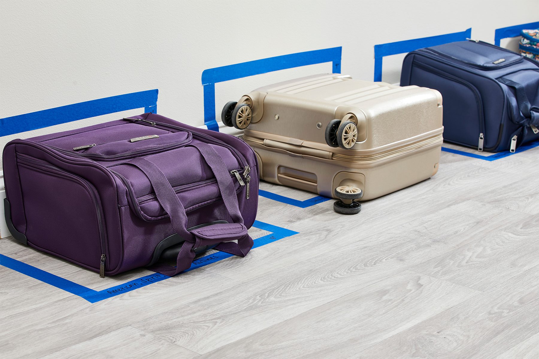 Suitcases rest against a wall, outlined by blue tape rectangles on a wood floor, with overhead lighting illuminating them.