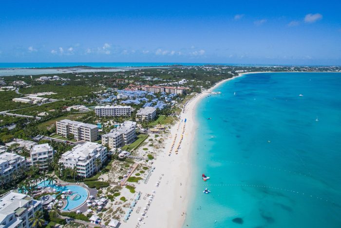 Beachfront hotels line a white sandy beach with turquoise water, under a clear blue sky, surrounded by lush greenery and distant buildings.