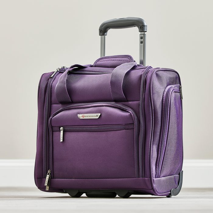 Purple wheeled suitcase stands on a light floor, featuring a retractable handle and multiple zippered compartments, against a neutral background.