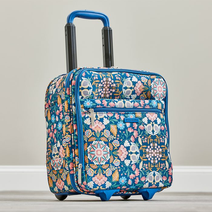 Wheeled floral suitcase stands upright, handles extended, against a light gray background; features colorful patterns and a small front pocket.