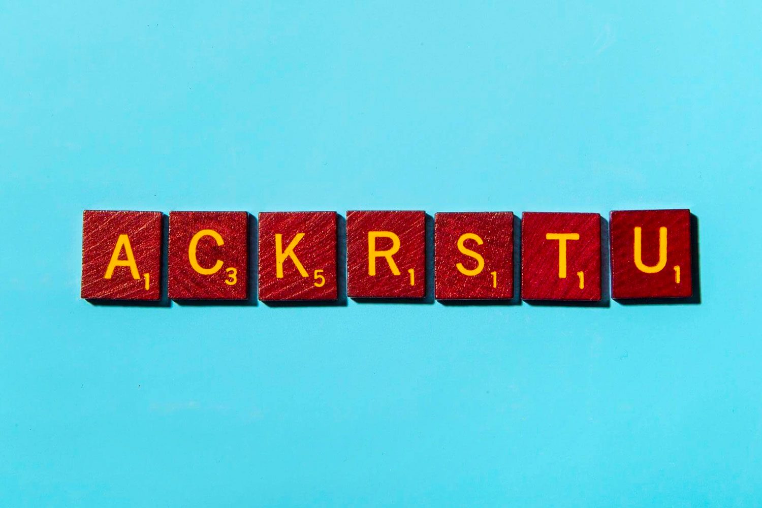 Scrabble tiles spell "ACKRSTU" on a bright blue background, with yellow letters and red tiles showing point values.