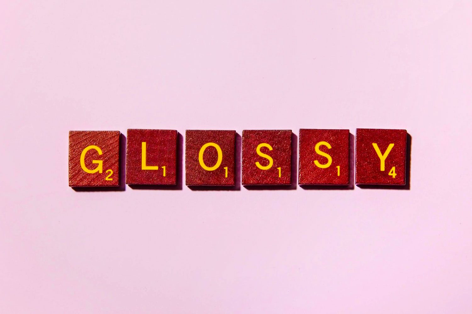 Scrabble tiles spell "GLOSSY" on a light pink background.